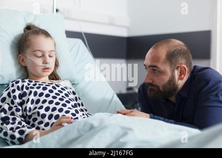 Thoughtful sad father looking at sleeping sick little daughter while in pediatric clinic patient room. Ill little girl resting after medical procedure while upset worried father sitting besides her. Stock Photo