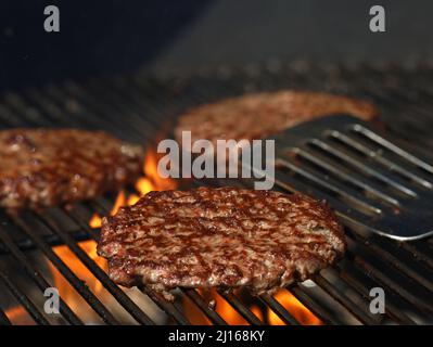 Freshly grilled beef burgers on grill grate with flames and stainless steel spatula in background Stock Photo