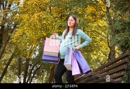 While walking down the street, pregnant woman start having uterus contractions. Mother with packages in her hands after shopping standing at the bench, holding her waist. Stock Photo