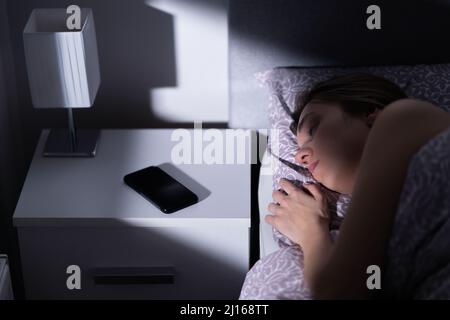 Sleeping woman in bed with phone on table at night. Cellphone on nightstand. Smartphone with silent mode, mute or turned off next to resting person. Stock Photo