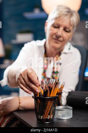 Focus on elderly woman retired artist hand picking a pencil for black and white professional sketch in home studio. Creative older person choosing drawing instrument from personal collection. Stock Photo