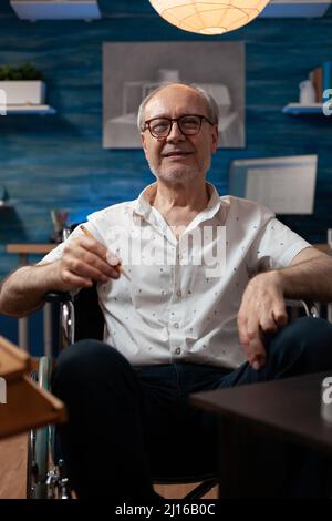 Portrait of senior man artist holding pencil sitting in wheelchair in front of easel with canvas in drawing workshop. Confident retired art teacher posing inspired in home professional studio. Stock Photo