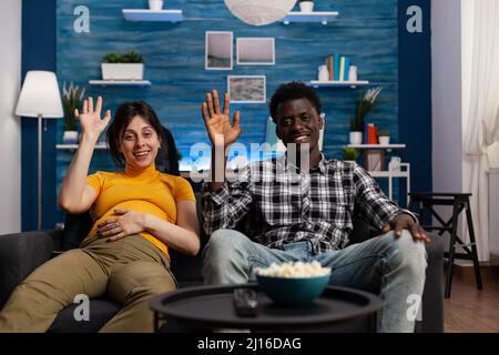 POV of happy multiethnic couple on videoconference call in living room. while expecting newborn. Young pregnant wife and smiling husband waving at video call while talking to relatives on internet. Stock Photo
