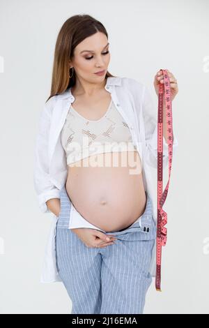 Portrait of young beautiful pregnant woman with long dark hair and makeup wearing white top and shirt, holding jeans.