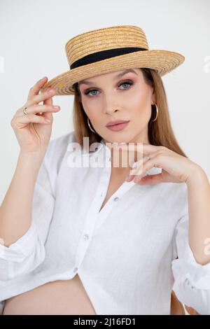Portrait of young blue-eyed pregnant woman with long dark hair and makeup wear golden earrings, white shirt, straw hat. Stock Photo