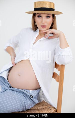Portrait of young pregnant woman with long dark hair and makeup wearing golden earrings, white shirt and straw hat.