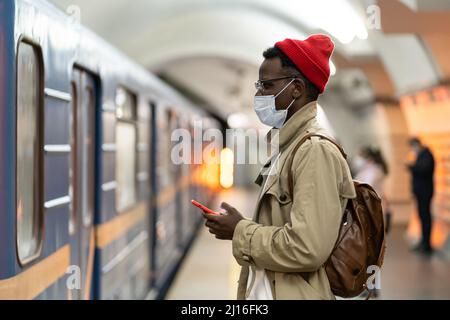 African man in face mask in subway station wait for train, using smartphone, scrolling social media Stock Photo
