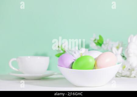 Easter eggs, pink, green and lilac, in a white plate, next to flowers with a cup of tea or coffee, on a green blue background. Pastel shades. Hard sha Stock Photo