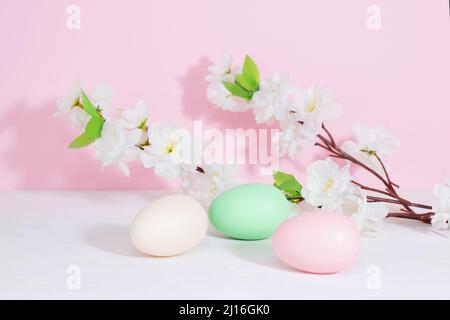 Easter eggs, pink, beige and green, on a concrete white table, next to flowers on a pink background. Pastel shades. Copy space Stock Photo