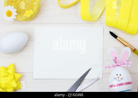 White notepad on a white wooden background. The Happy Easter Bunny is created from an egg. The moment of creating an Easter decoration. top view, flat Stock Photo