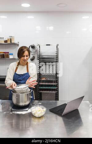 Female pastry chef using a laptop computer while making tasty baking in the kitchen. Stock Photo