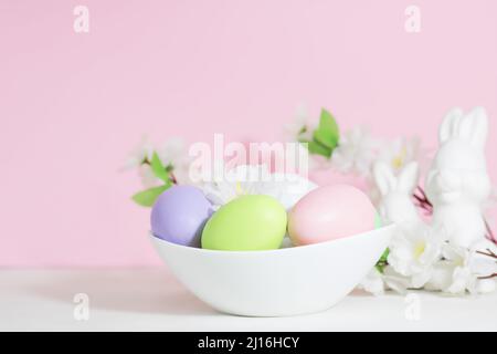 Easter eggs, pink, green and lilac, in a white plate, near rabbits and flowers on a pink background. Pastel shades. Stock Photo
