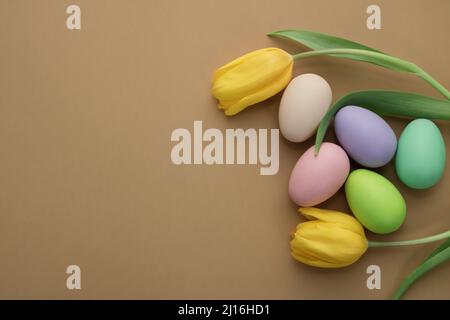 Beige background with green, pink, light green, lilac, white and beige Easter eggs and beautiful yellow tulips. Colorful Easter eggs. View from above. Stock Photo