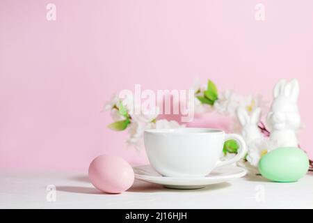 White mug with tea or coffee near pink and green Easter eggs, rabbits and flowers, on a concrete white table, on a pink background, with hard shadows. Stock Photo