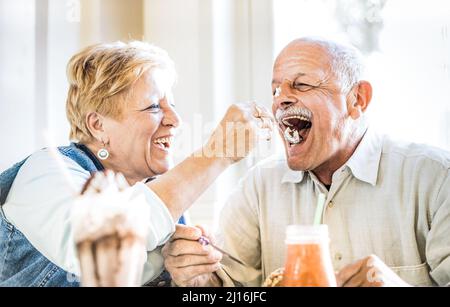 Happy retired senior couple in love enjoying bio icecream cup - Joyful elderly lifestyle concept - Wife feeding husband and having fun at bar cafe res Stock Photo