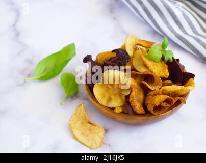 Vegan snacks, multicolored vegetable chips in wooden bowl and set of fresh farmer vegetables, rustic still life, selective focus. Stock Photo