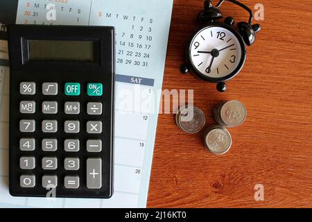 Top view image of calculator, calendar, alarm clock and coins on wooden table. Stock Photo