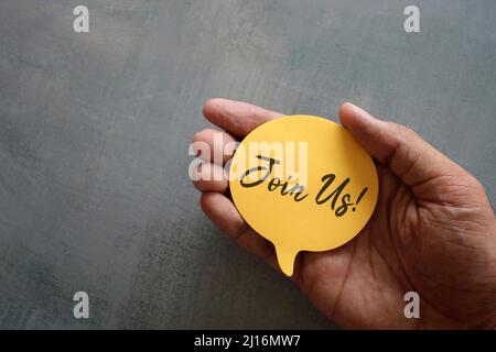 Hand holding yellow speech bubble with text JOIN US! with copy space. Registration and hiring. Stock Photo