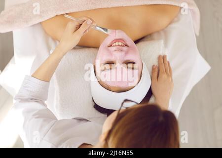 Unknown female beautician with brush applies pink mask on face of satisfied client. Stock Photo