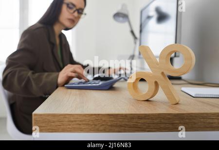 Close up of wooden percent sign standing on table on background of woman counting on calculator. Stock Photo