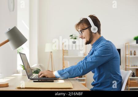 Young happy man in headphones typing on laptop and using internet web online services. Stock Photo