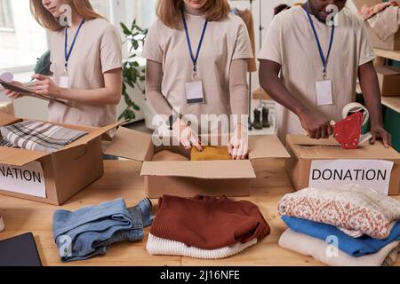 High angle shot of three unrecognizable volunteers wearing beige T-shirts packing clothes into cardboard boxes for donation Stock Photo