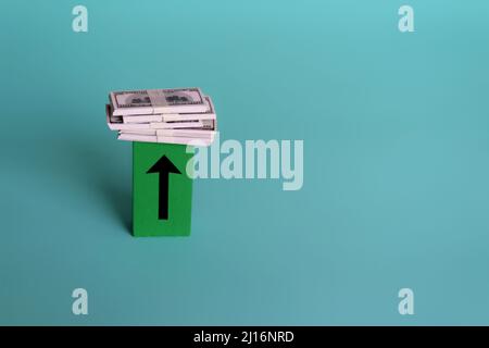 Pile of money on top of wooden cube with upward arrow. Copy space. Boost income, profit, surplus. Stock Photo