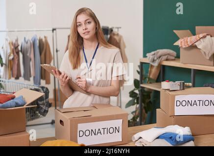 Modern young woman with long hair working in clothes charity organization making list of donation boxes looking at camera Stock Photo