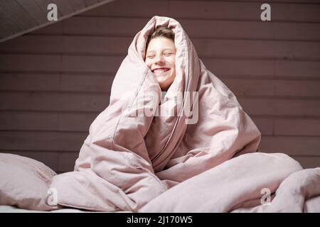 Boy teen sitting the bedroom under the pink blanket over head. Smiling face in the morning Stock Photo