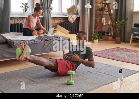 Horizontal shot of sporty young African American man wearing comfortable clothes doing morning exercise at home using dumbbell Stock Photo