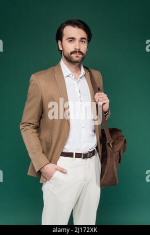 man with mustache standing with hand in pocket and holding leather bag on dark green Stock Photo