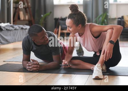 Young woman sitting on floor chatting to her boyfriend while he is doing plank exercise in morning Stock Photo