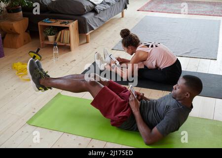 Horizontal high angle view shot of young Black man and his Caucasian girlfriend doing morning workout at home and watching something on smartphones Stock Photo