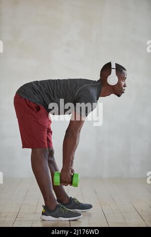 Vertical side view shot of young African American man exercising with use of dumbbells and listening to music in headphones Stock Photo