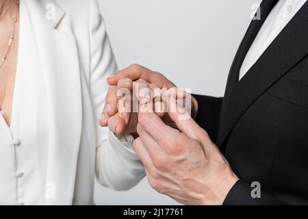 Cropped view of groom wearing ring on finger of bride in formal wear isolated on grey Stock Photo