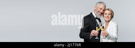 Positive mature couple in wedding clothes holding champagne and looking at camera isolated on grey, banner Stock Photo