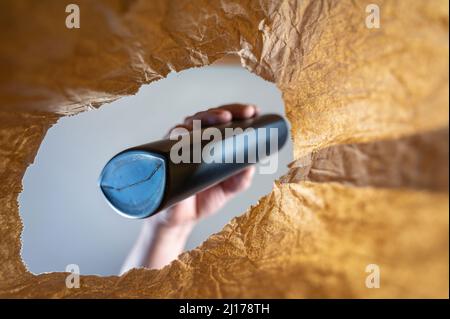A hand pulls a black plastic bottle from a paper bag. A mature man's hand holds the shampoo bottle over the open brown bag. Shot from the bottom up. C Stock Photo