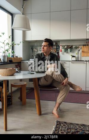 Thoughtful young man sitting at table in kitchen Stock Photo