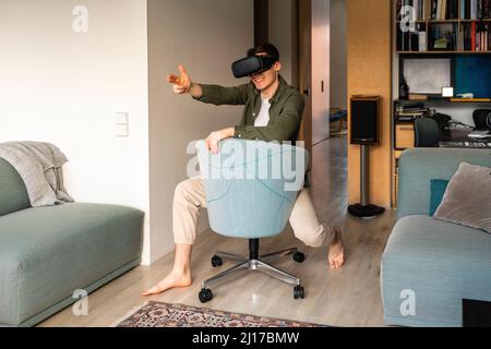 Smiling young man wearing virtual reality simulator gesturing on chair at home Stock Photo