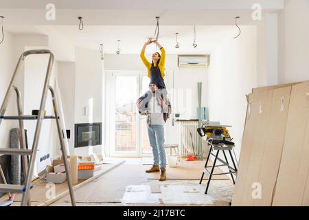 Young man carrying girlfriend on shoulders installing light bulb in living room Stock Photo