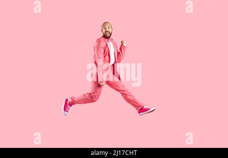 Happy excited funny young man in suit and sneakers having fun jumping on pink background. Stock Photo