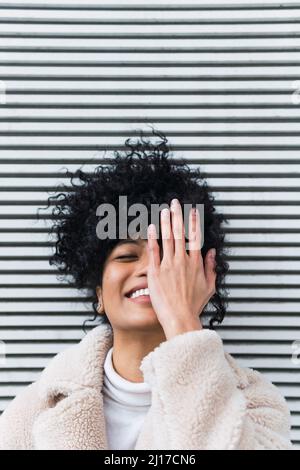 Smiling young woman covering face in front of wall Stock Photo