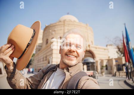 Happy Senior man traveler with hat and backpack make selfie photo sun day. Concept travel older male lifestyle antique. Stock Photo