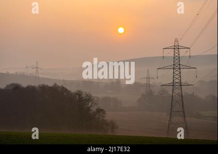 Ledgerwood in Scottish Borders, UK. 23rd March 2022. Engery, power, electricity. The sun rises on another day of high energy prices as the cost of living continues to rise. The transition to green energy is high on the agenda but also the balance of reducing costs of energy currently high due to the covid pandemic and the current war in Ukraine and sanctions against Russia. The view is of electricity pylons and a distance wind turbine as the sun rises near Ledgerwood in the Scottish Borders. Credit: phil wilkinson/Alamy Live News Stock Photo