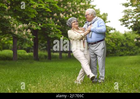 Senior man and woman are dancing on the green grass in the park among the trees. Stock Photo