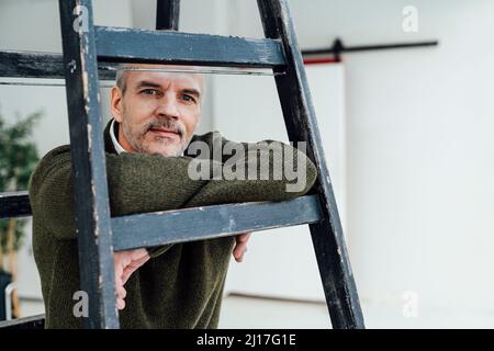 Confident businessman leaning on ladder in office Stock Photo