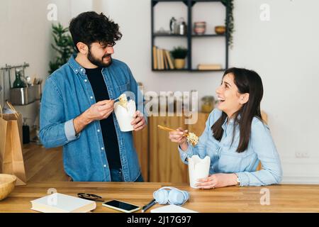 Smiling woman and man eating noodles with chopsticks in kitchen at home Stock Photo