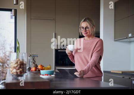 Blond woman holding coffee cup using tablet PC in kitchen Stock Photo