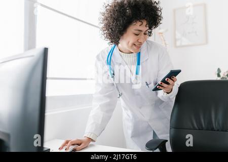 Smiling doctor with curly hair using smart phone at clinic Stock Photo