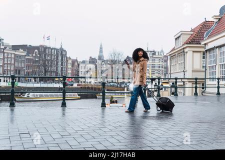 Woman with smart phone pulling wheeled luggage walking on footpath in city Stock Photo